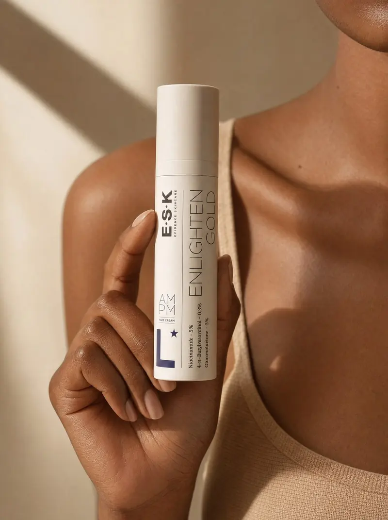 A woman with deep-toned skin holding ESK Enlighten Gold face cream in warm studio light